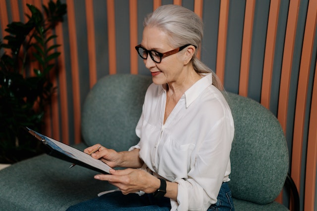 Woman in White Dress Shirt and Black Pants Wearing Black Framed Eyeglasses Sitting on Gray Couch